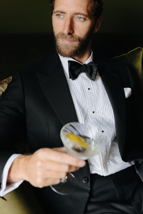 Model wearing white pleated budd dress shirt with black bow tie, sat on the sofa drinking a martini