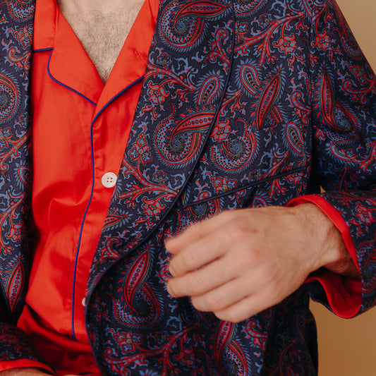 Close up of male model sat on a chair wearing Men's Fine Twill Cotton Pyjamas in Cardinal Red with madder silk paisley dressing gown on a plain cream background